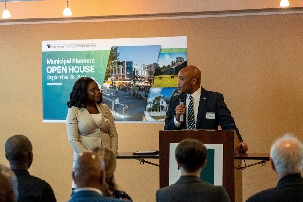 Two people stand and speak at a podium during a Municipal Planners Open House event. A sign behind them displays event details and photos. Several attendees are seated and listening in the foreground.