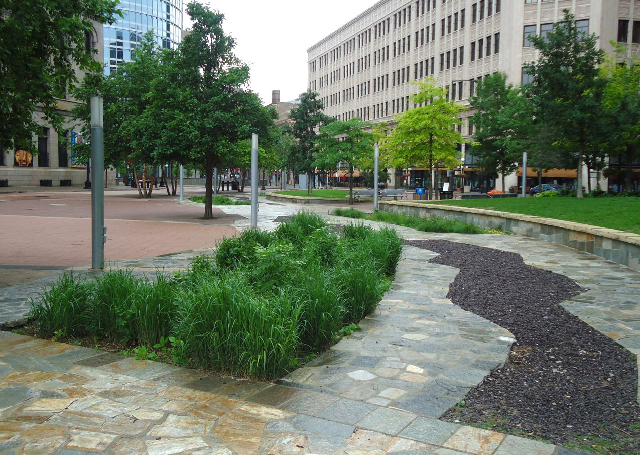 A landscaped urban plaza features trees, green plants, and a winding stone path bordered by a dry creek bed, with tall office buildings surrounding the area.