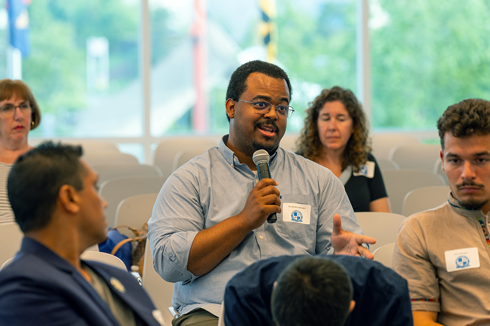 A man holding a microphone speaks during a discussion in a room with several seated people. The background shows windows with trees outside, and attendees appear attentive and engaged.