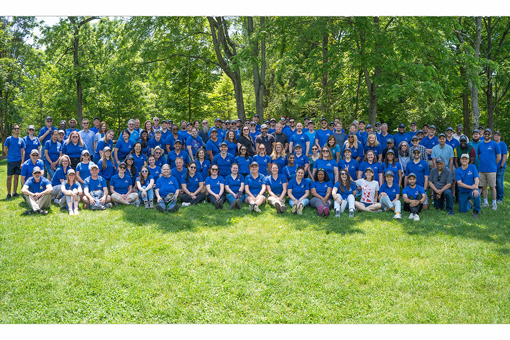 A large group of people wearing matching blue shirts pose together outdoors on a grassy area, surrounded by green trees under bright daylight.