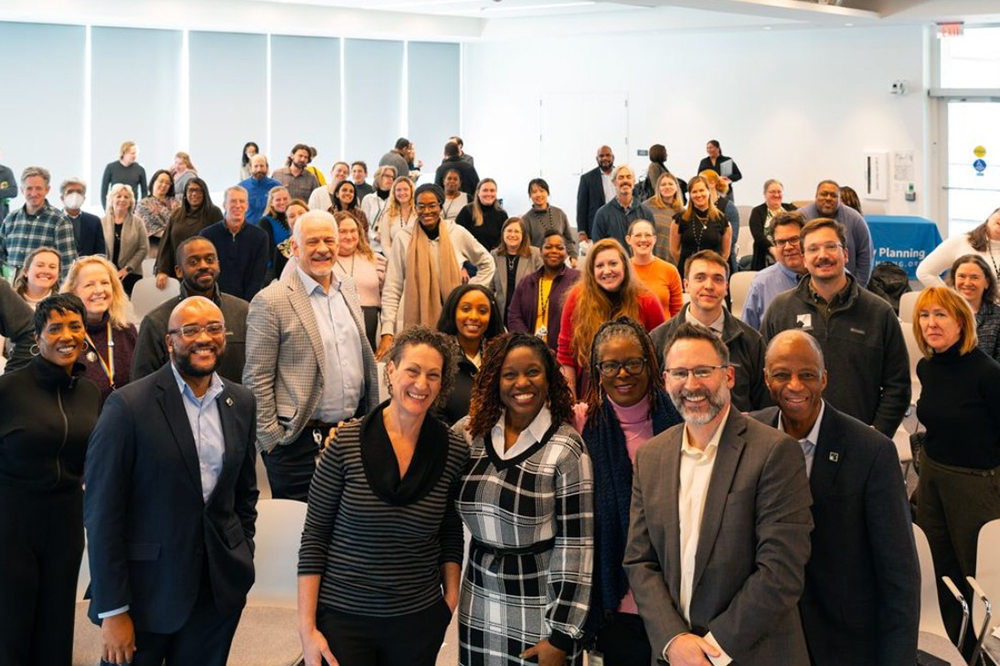 A large, diverse group of adults stands and smiles at the camera in a bright, modern room with white walls and large windows. Some people are dressed in business attire, and the atmosphere appears friendly and welcoming.