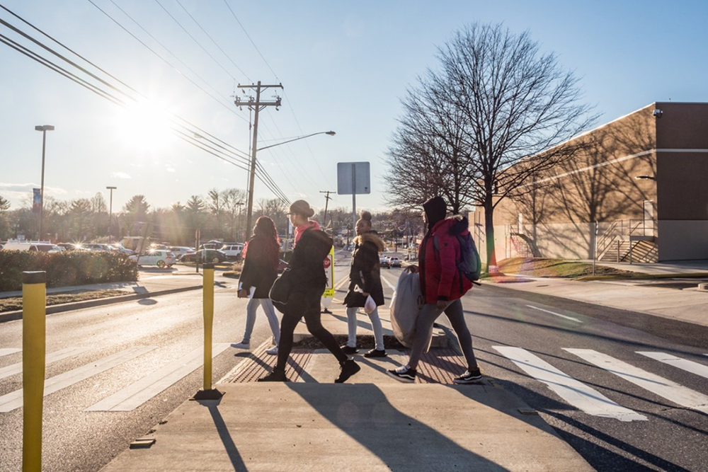 Five people walk across a crosswalk in bright sunlight near a parking lot and a large building, with bare trees and streetlights in the background.