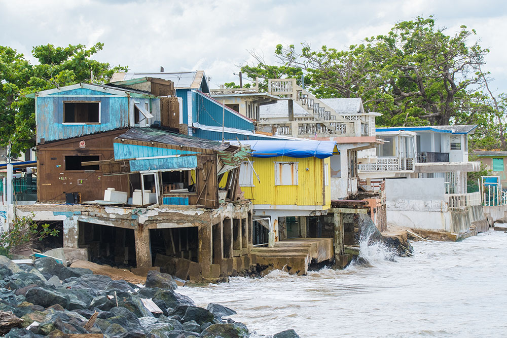 Colorful, weathered houses built on stilts stand along a rocky shoreline, with waves crashing against their foundations; some buildings show visible damage and erosion from the sea. Trees and greenery are in the background.