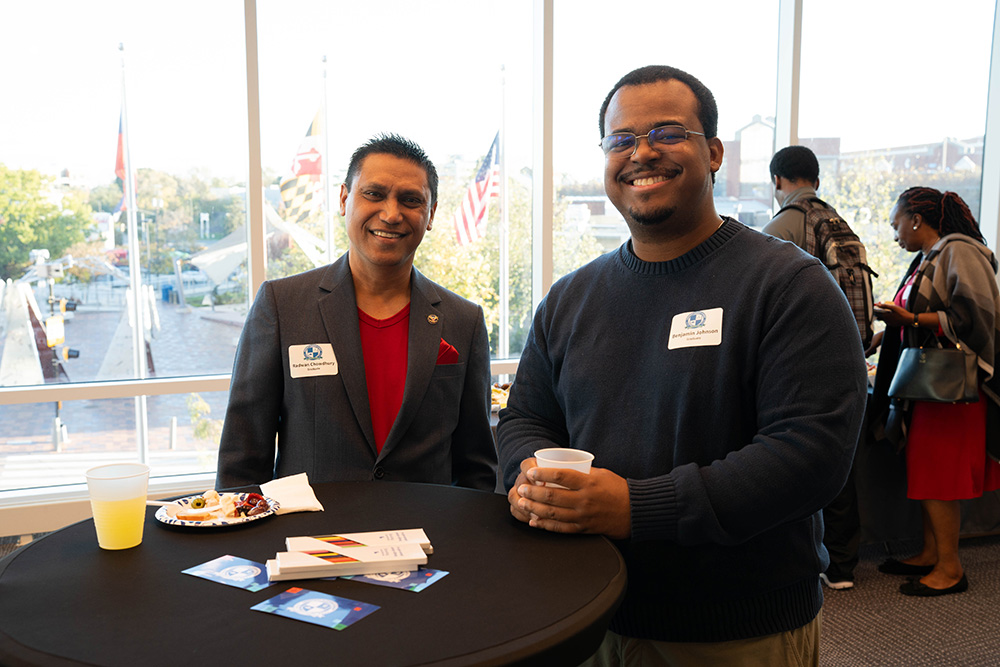 Two men stand and smile at a round table with drinks and snacks at a professional event. Name tags and business cards are visible. Large windows in the background show flags and trees outside.