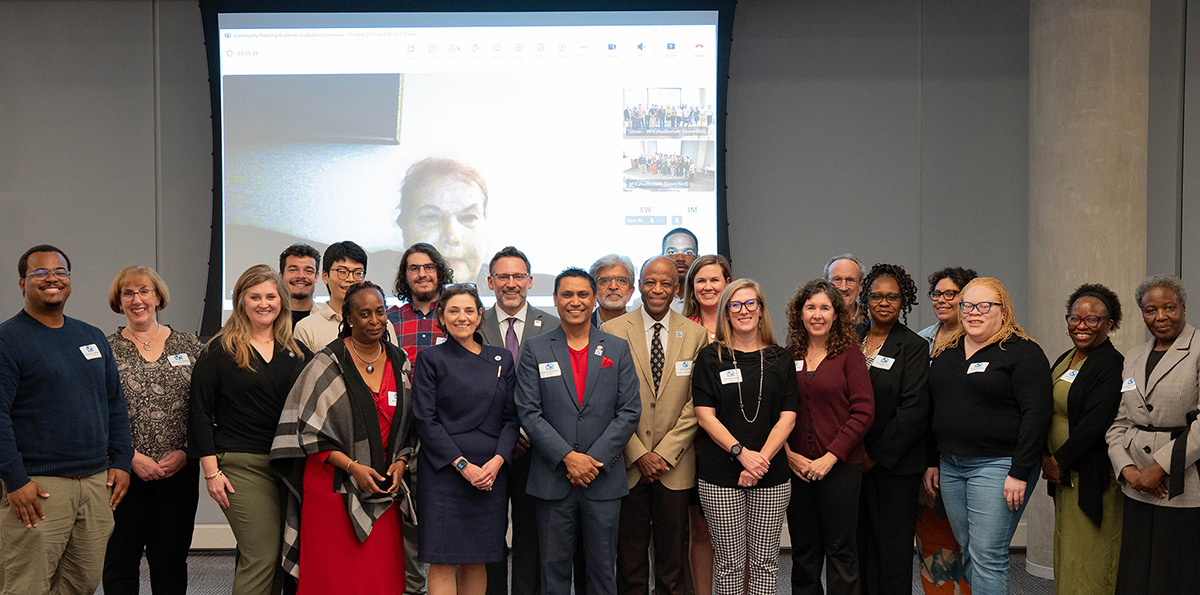 A diverse group of professionals poses and smiles for a group photo in front of a large screen displaying a video call with additional participants. The setting appears to be a conference or meeting room.