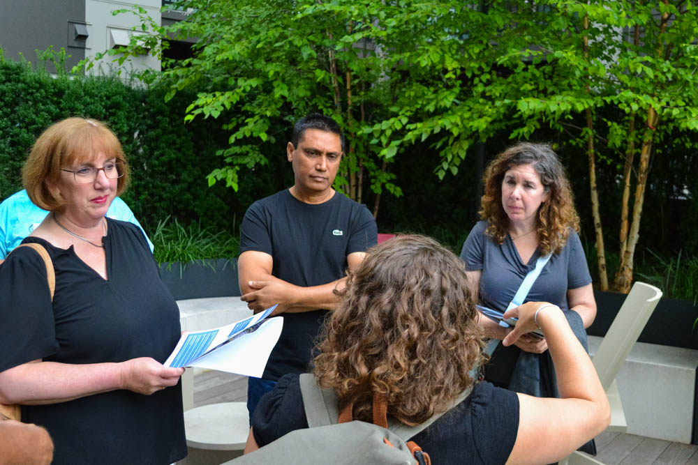 Four adults stand outdoors in discussion. Two women hold papers, one man crosses his arms, and another woman faces away from the camera. Green trees and bushes are in the background.