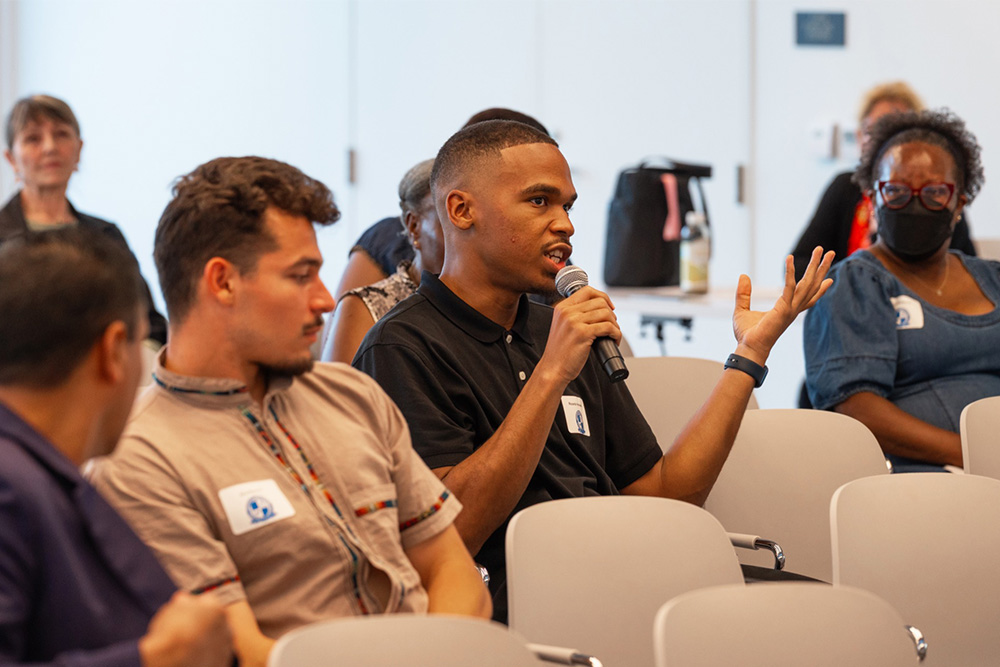 A young man speaks into a microphone and gestures with his hand while seated among others at an indoor event. People around him listen attentively; some are wearing name tags and one person wears a face mask.
