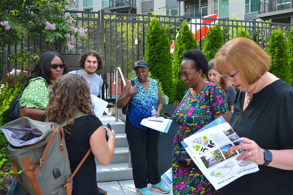 A group of people stand outdoors by a staircase, holding papers and clipboards, engaged in conversation. They appear to be on a tour or discussing plans, with greenery and an apartment building in the background.