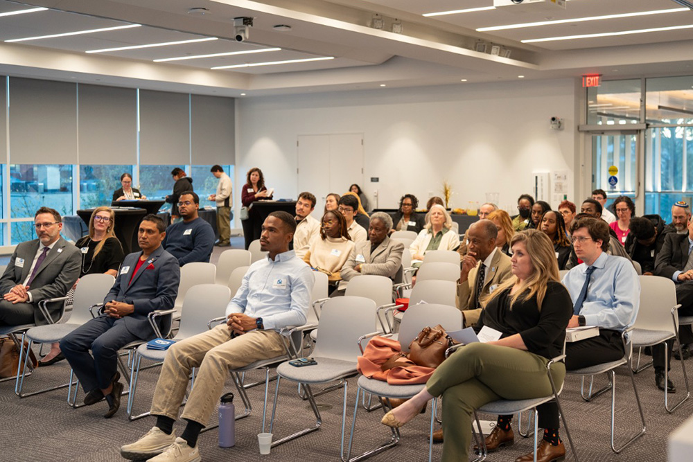 A diverse group of people sit attentively in a modern conference room, listening to a presentation. Some take notes, while others look forward. The room has large windows and rows of empty and occupied chairs.
