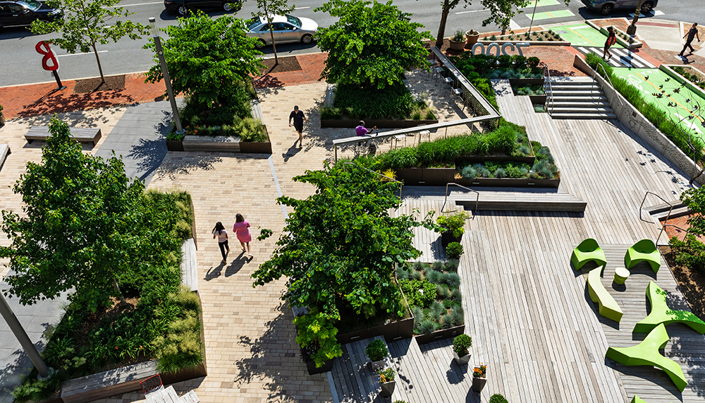 Aerial view of an urban park with wooden walkways, green trees, landscaped plants, people walking, unique green benches, and cars driving on adjacent streets. Steps and bike lanes are visible on the right.