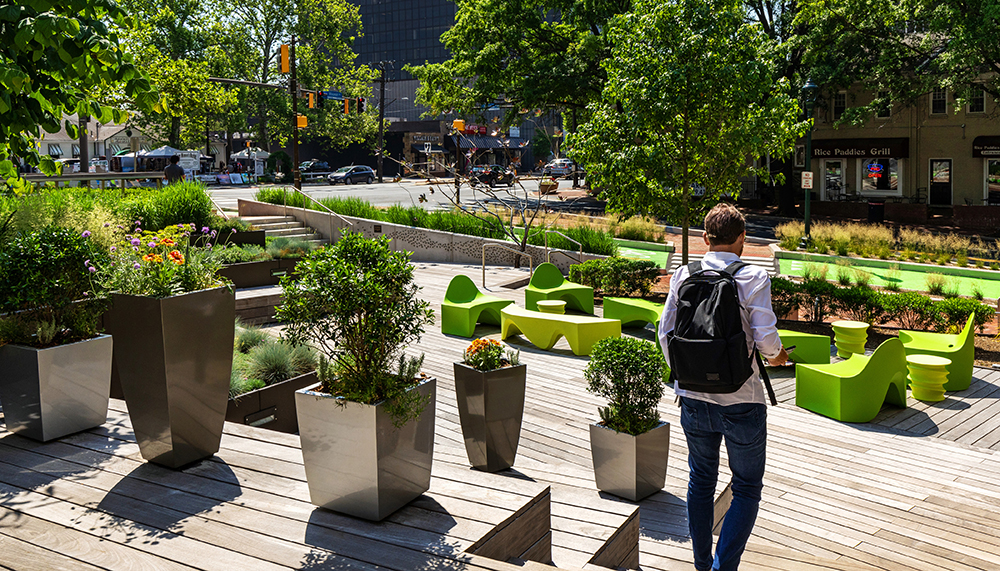 A person with a backpack walks on a wooden deck surrounded by modern planters filled with greenery and vibrant flowers, with bright green outdoor seating and trees nearby in an urban park setting.