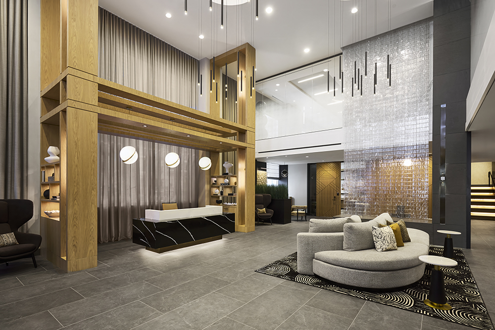 Modern hotel lobby with a sleek black and white reception desk, tall wooden beams, contemporary lighting, gray tile floor, curved gray sofas, and decorative wall tiles, creating a stylish and welcoming atmosphere.