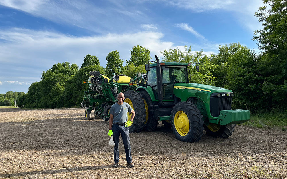 A person wearing gloves stands smiling in front of a large green tractor and farming equipment in a field, with trees and a blue sky with clouds in the background.