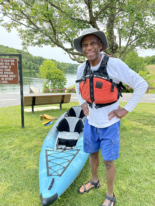 A man wearing a hat, life jacket, and blue shorts stands smiling beside an inflatable kayak on grass near a lake, with trees, a bench, and a sign visible in the background.