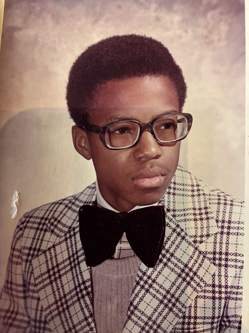 A young man with an afro hairstyle wears large glasses, a plaid jacket, a light sweater, and a black bow tie, posing for a portrait against a neutral background.