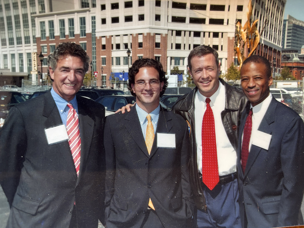 Four men dressed in business attire stand smiling together outdoors in a city setting with buildings, parked cars, and a construction site in the background. They are wearing name tags and appear to be at a formal event.