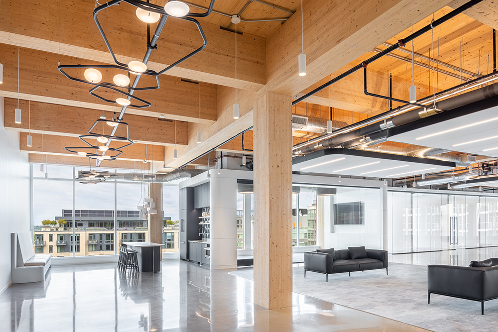 Modern open office space with large windows, exposed wooden beams, polished floors, geometric light fixtures, black sofas, a table with chairs, and plenty of natural light.