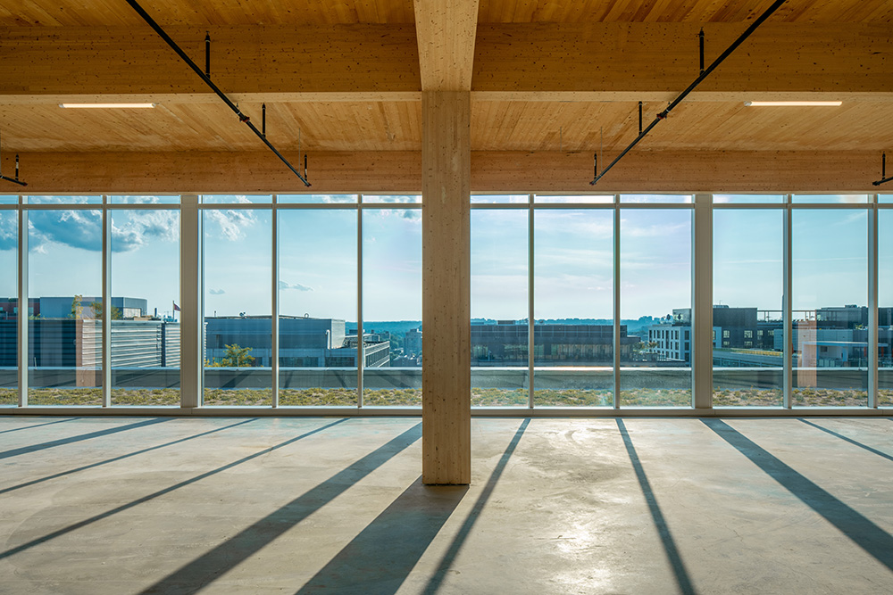 Spacious empty room with concrete floors and large wooden beams, featuring floor-to-ceiling windows that overlook a cityscape and let in ample natural light. Shadows from the structure are cast across the floor.