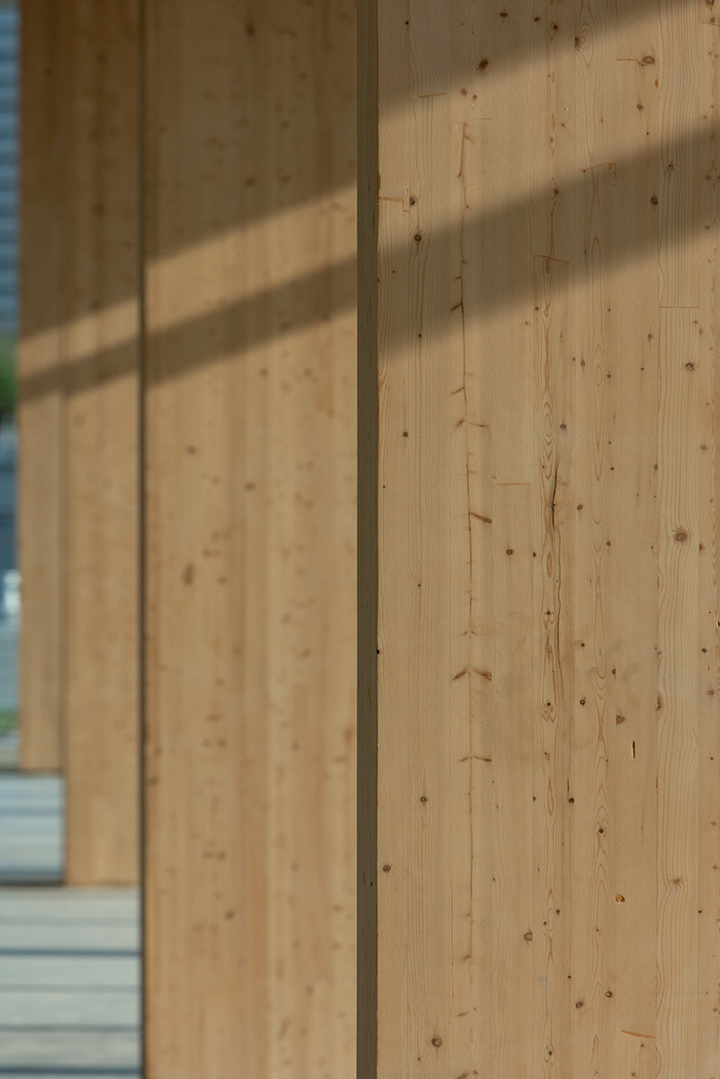 Close-up view of vertical wooden panels with visible grain and knots, casting soft shadows in natural light. The scene suggests an architectural or construction detail, with a modern, minimalistic style.