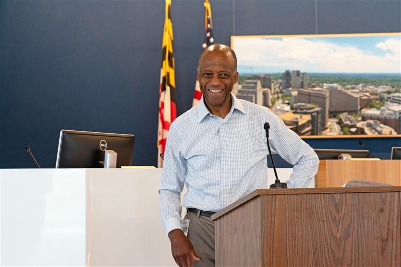 A man in a light blue shirt stands smiling at a podium in an office or conference room, with Maryland and U.S. flags and a cityscape photo in the background.