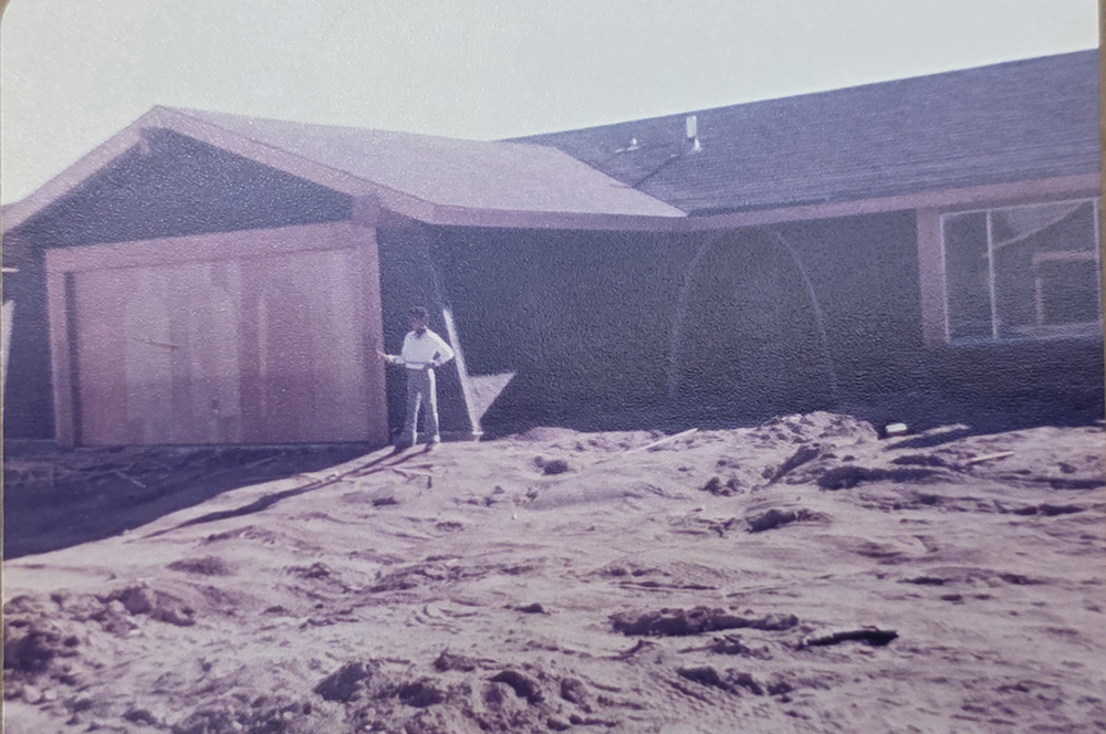 A person stands in front of a partially constructed house with exposed wood and a dirt yard. The garage door is not yet installed, and the ground appears uneven and unfinished.