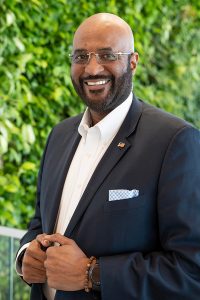 A smiling man in a suit and glasses stands in front of a leafy green background, holding his suit jacket with both hands. He wears a white shirt, checkered pocket square, and a beaded bracelet.