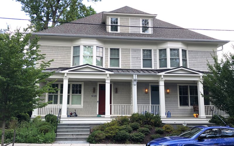 A two-story gray house with a wide front porch, white trim, and dormer windows. There are bushes and a green lawn in front, with a blue car parked on the street.