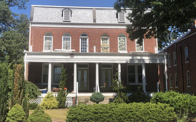 A two-story red brick building with white columns and trim, arched windows on the second floor, and a covered front porch. Well-maintained landscaping with shrubs and small trees is visible in front.