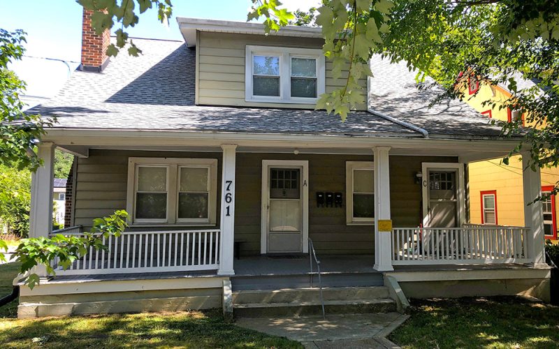 A two-story house with light gray siding, white trim, a covered front porch with white railings, and the number 761 on the post. Trees provide shade, and a concrete path leads to the front steps.