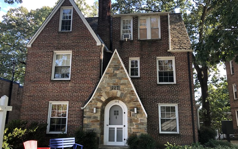 A three-story brick house with a steep roof, central stone entryway marked "721," and symmetrical windows. A red chair and a blue bench sit on the lawn near the entrance, surrounded by trees.