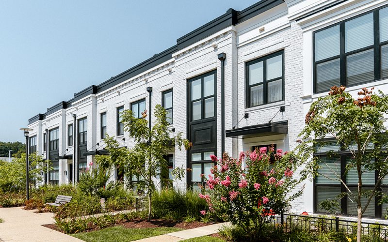 A row of modern white townhouses with black trim, large windows, and colorful landscaping, including flowering bushes and young trees, along a clean sidewalk under a clear sky.