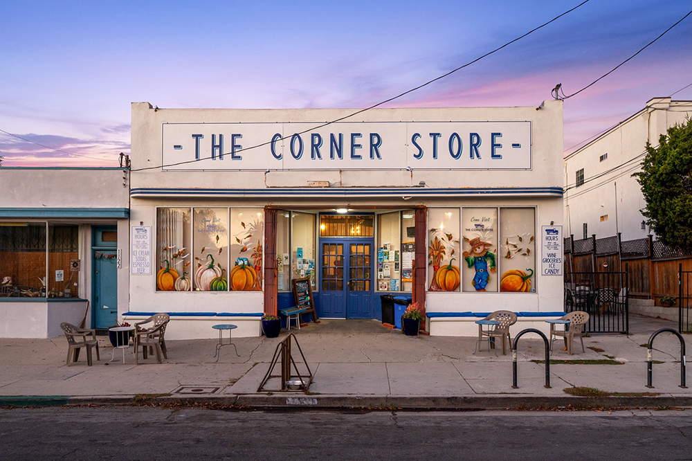A small white storefront labeled "The Corner Store" with blue trim, large painted pumpkins and flowers on the windows, outdoor chairs and tables, and a glowing sunset sky in the background.
