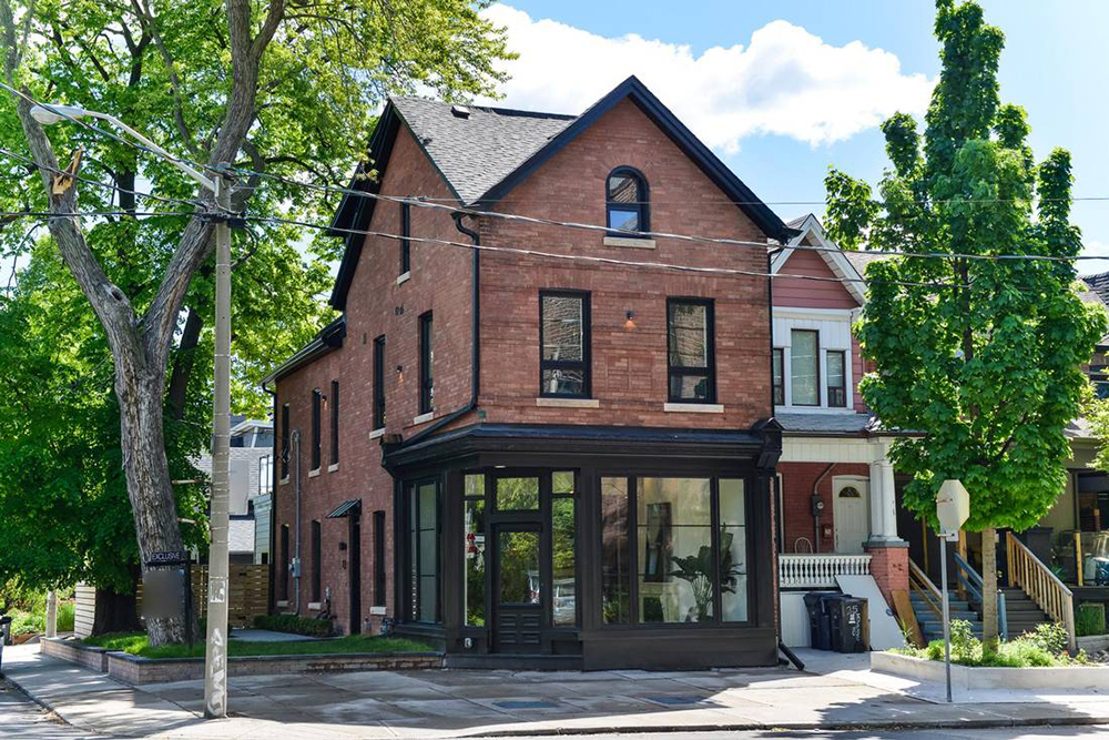 A red-brick corner building with large ground-floor windows and black trim, next to a leafy tree and neighboring houses, under a bright blue sky with scattered clouds.