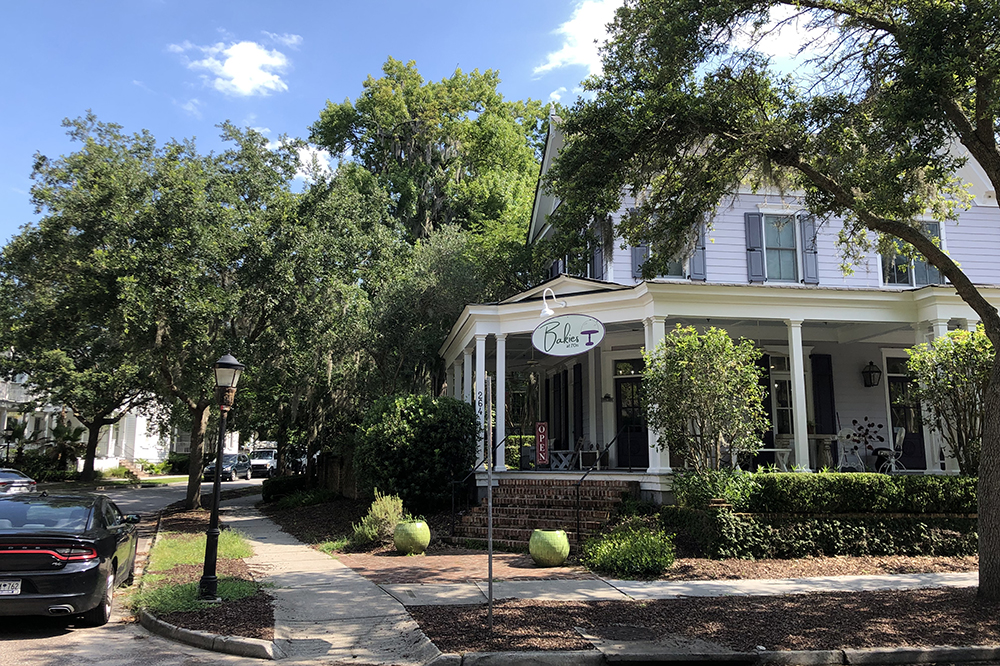 A corner view of a tree-lined residential street with a white house featuring a wraparound porch and a sign reading "Blake T". There are large potted plants, greenery, and a parked black car nearby.