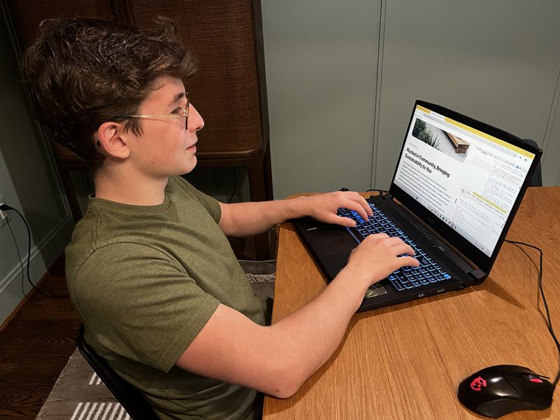 A young person wearing glasses and a green t-shirt types on a laptop at a wooden table. The screen displays a webpage about sustainable building design. A black mouse is next to the laptop.