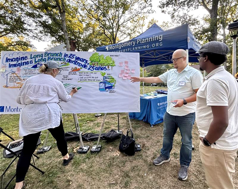Three people stand by a large illustrated board under a blue Montgomery Planning tent at an outdoor event. One person writes on the board while the others discuss, surrounded by trees and grass.