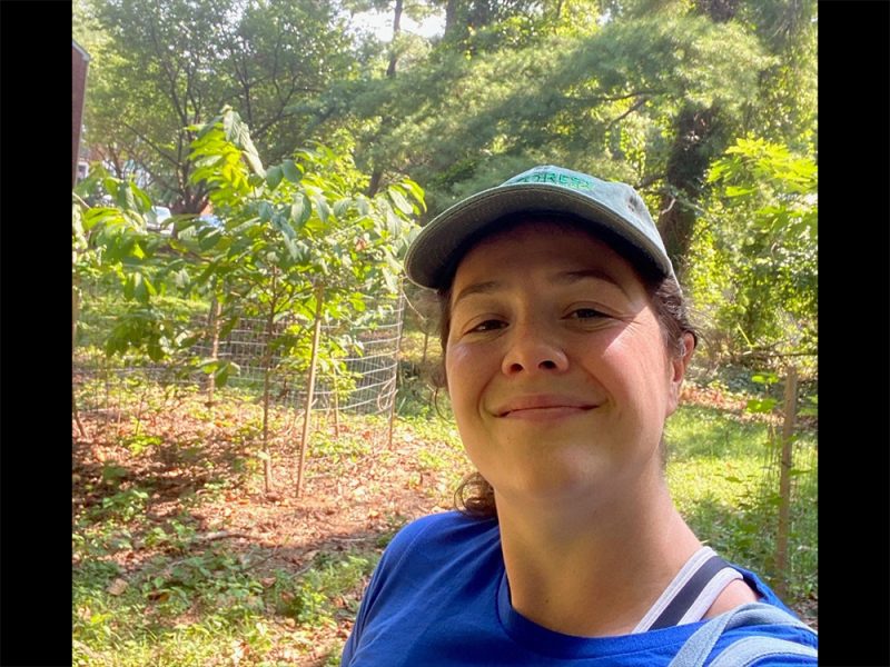 A person wearing a blue shirt and a light green cap smiles while standing outdoors in a sunny, green garden with young trees and wire fencing visible in the background.