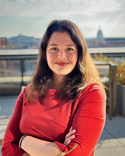 Kelsey Desmond stands in a brightly colored shirt, smiling, with their arms crossed with the U.S. Capitol visible in the background.