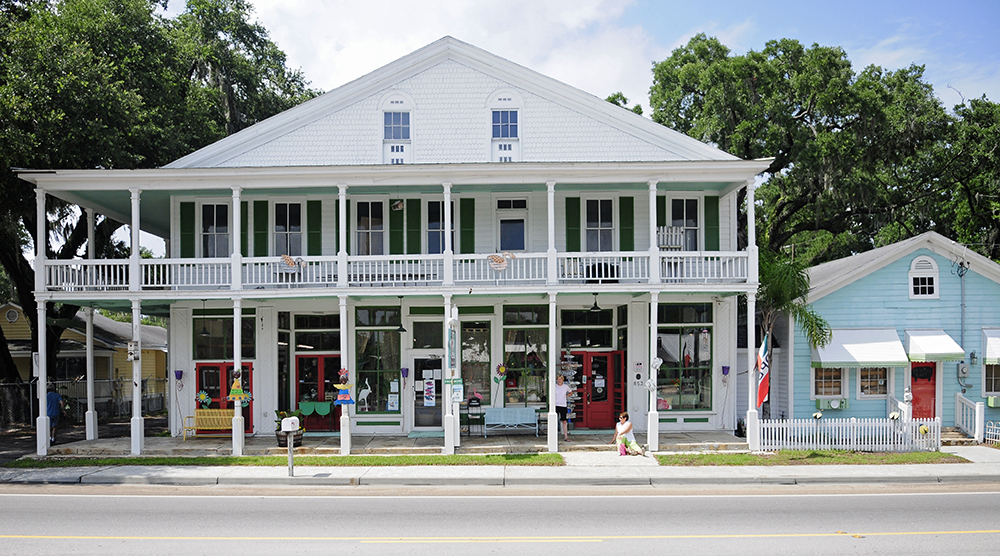 A two-story white historic building with a wraparound balcony sits beside a small blue house with a white picket fence, both facing a street and surrounded by large green trees under a partly cloudy sky.