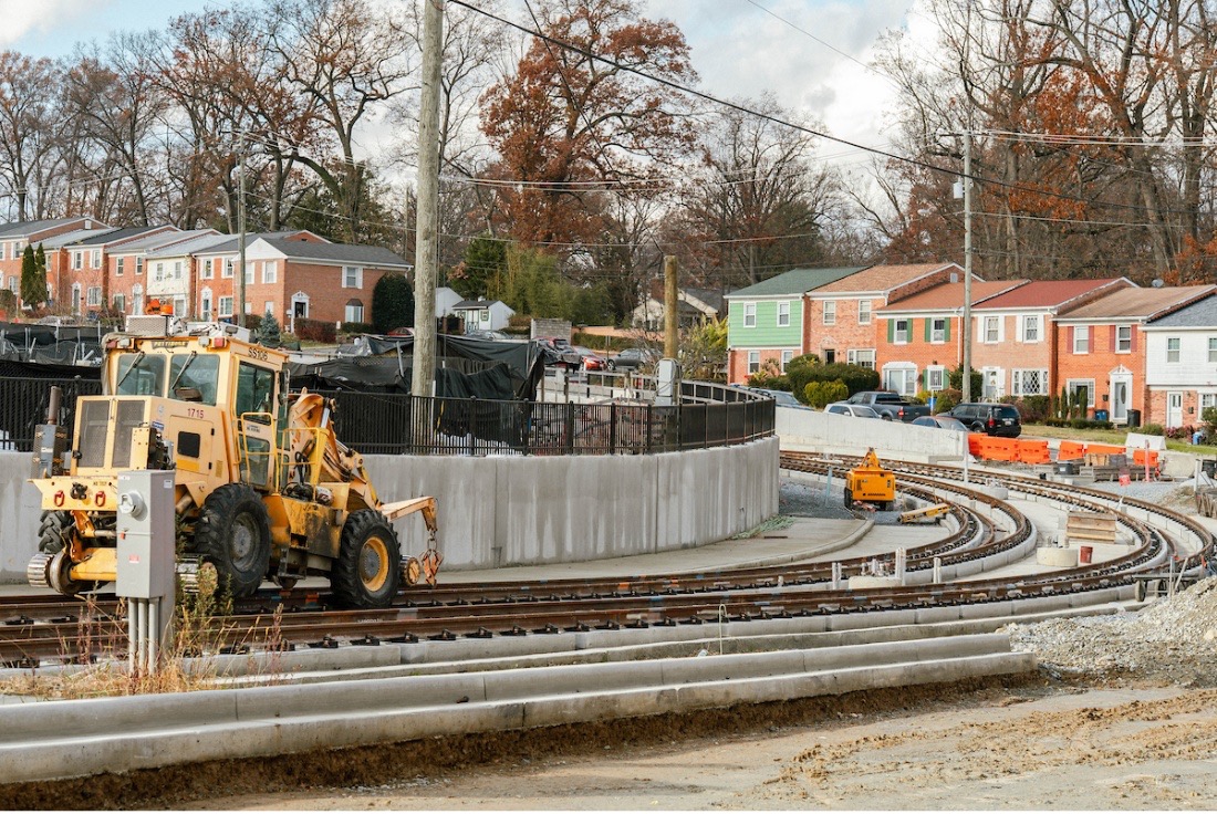 A construction vehicle works near curved light rail tracks in a residential neighborhood with rows of brick townhouses and trees in the background.