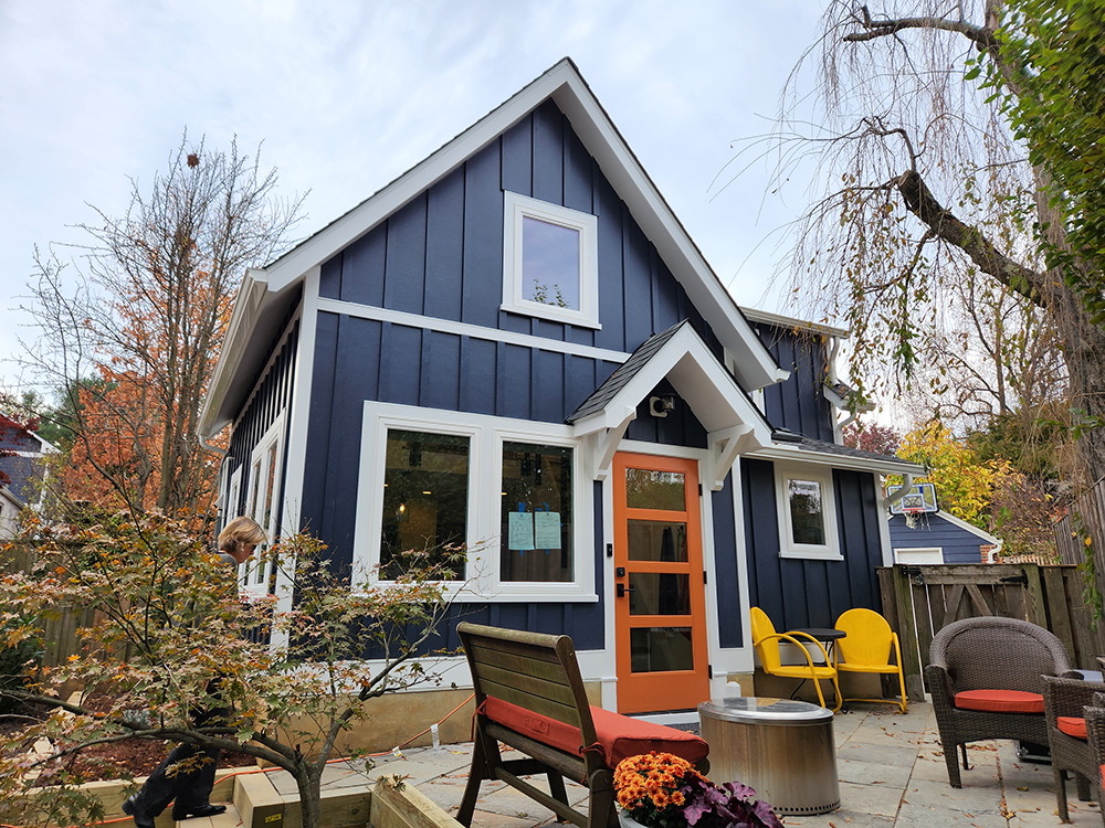 A modern blue house with white trim and an orange door, featuring large windows. The patio has outdoor furniture, including wooden benches and yellow chairs, surrounded by trees with autumn leaves.