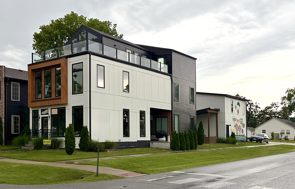Modern three-story house with large windows and a rooftop deck, located on a corner lot with well-kept grass and shrubs; neighboring homes and a cloudy sky in the background.