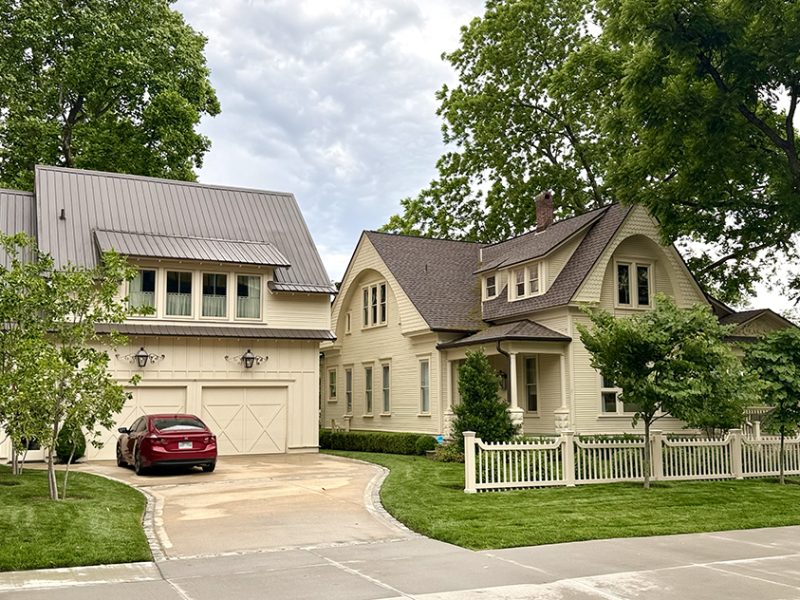 A cream-colored house with a detached garage, a red car parked in the driveway, white picket fence, manicured lawn, and large green trees surrounding the property under a cloudy sky.