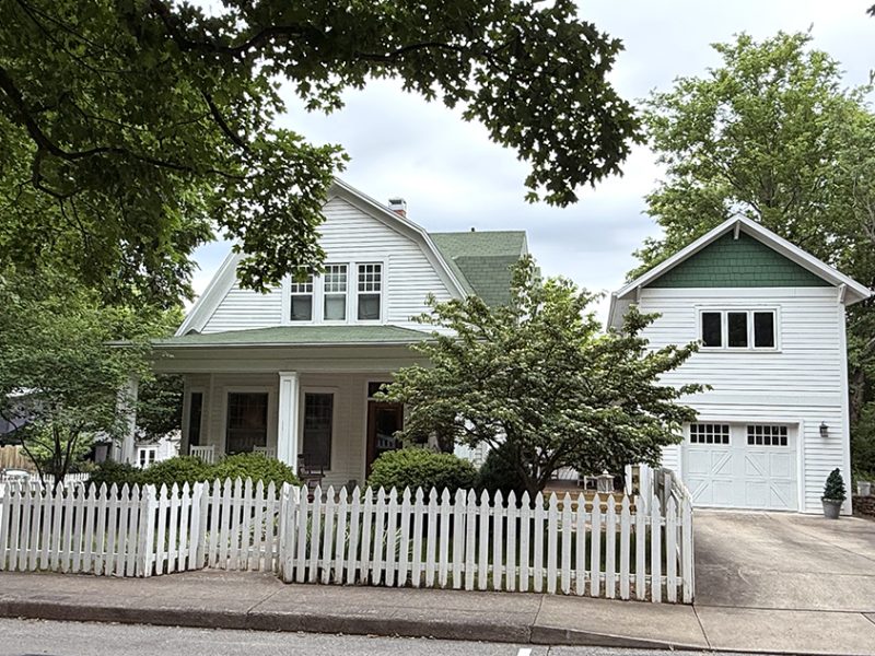 A white two-story house with green roof accents and an attached garage sits behind a white picket fence. Trees and shrubs surround the house, and a concrete driveway leads to the garage.