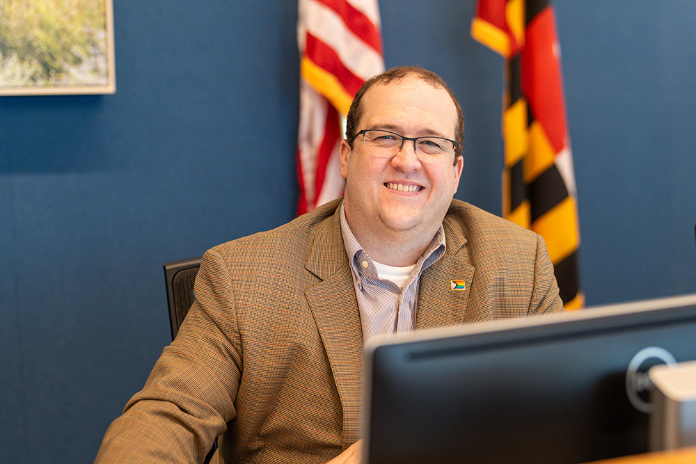 A man wearing glasses and a brown plaid blazer sits at a desk, smiling at the camera. American and Maryland state flags are visible in the background.