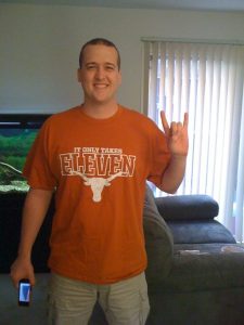 A smiling man wearing a burnt orange "It Only Takes Eleven" Texas Longhorns shirt stands indoors, holding up a "hook 'em horns" hand sign and a phone, with a couch and vertical blinds in the background.