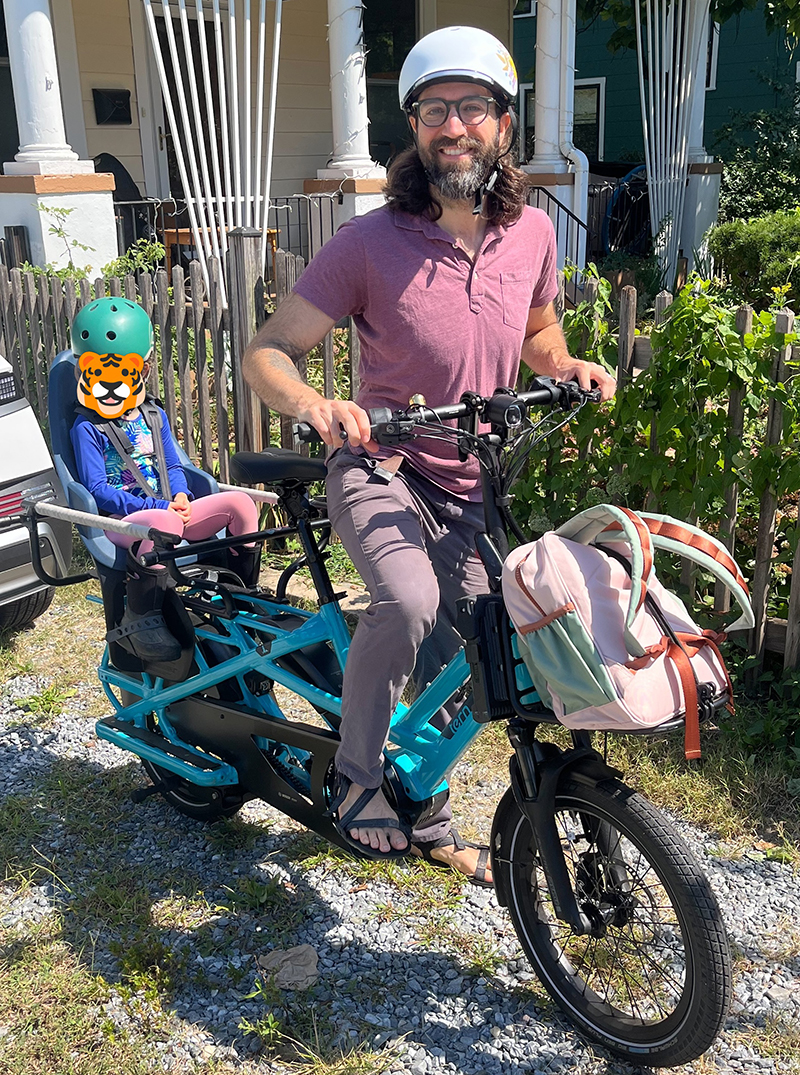 A smiling man wearing a white helmet sits on a blue electric cargo bike with a pink-and-green backpack on the front; a child with a tiger-face sticker and helmet sits in the rear child seat.