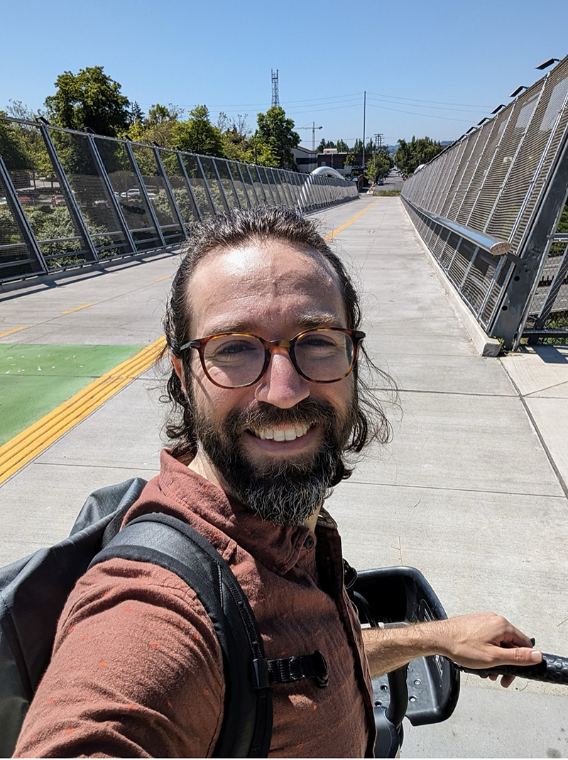 A man with glasses and a backpack smiles for a selfie on a sunny day, standing on a pedestrian bridge with a fence on both sides and trees in the background.