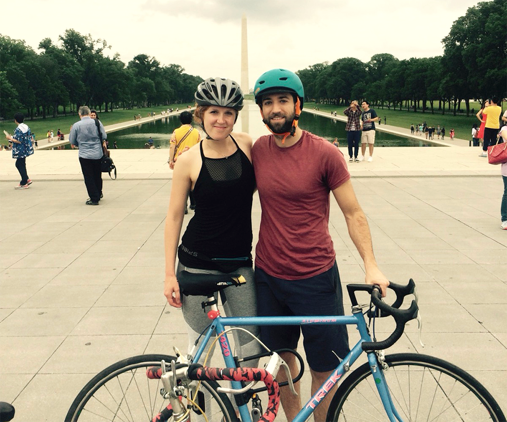 A woman and a man with helmets pose smiling beside a blue bicycle in front of the Lincoln Memorial Reflecting Pool, with the Washington Monument visible in the background. Other people are walking around.