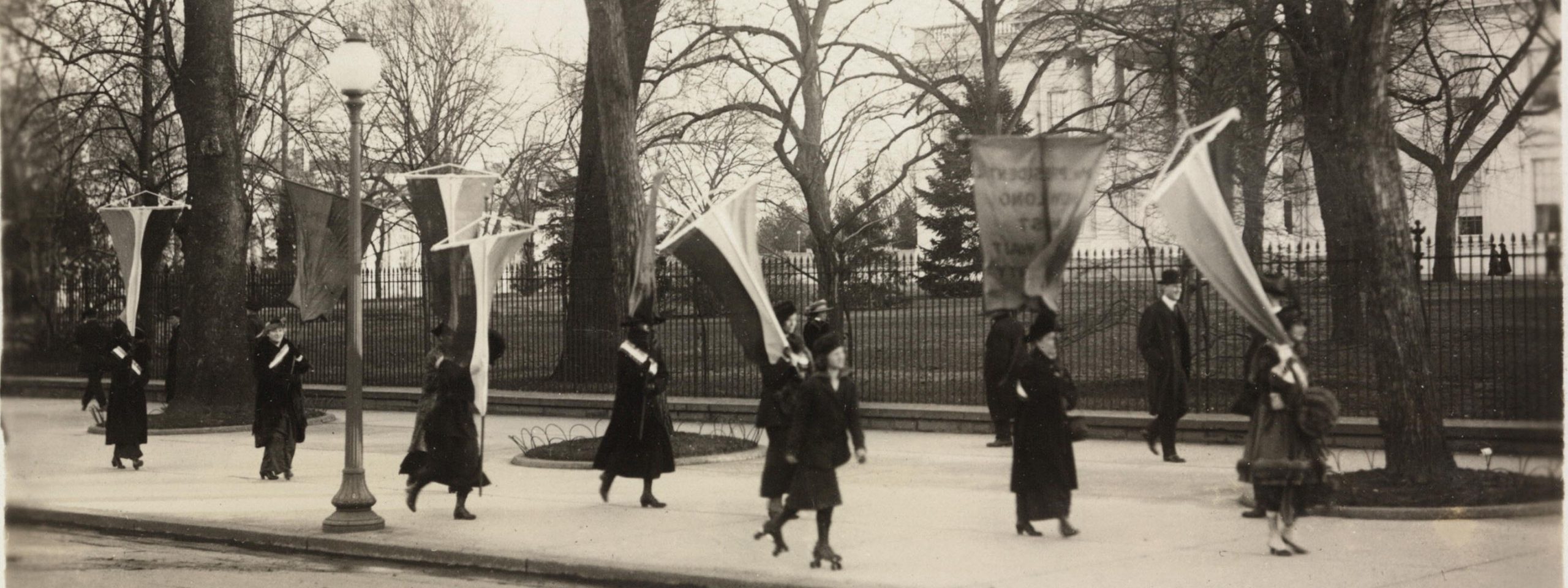 Women walk past the Whie House holding pro-suffrage flags and banners. One woman passes on roller skates.